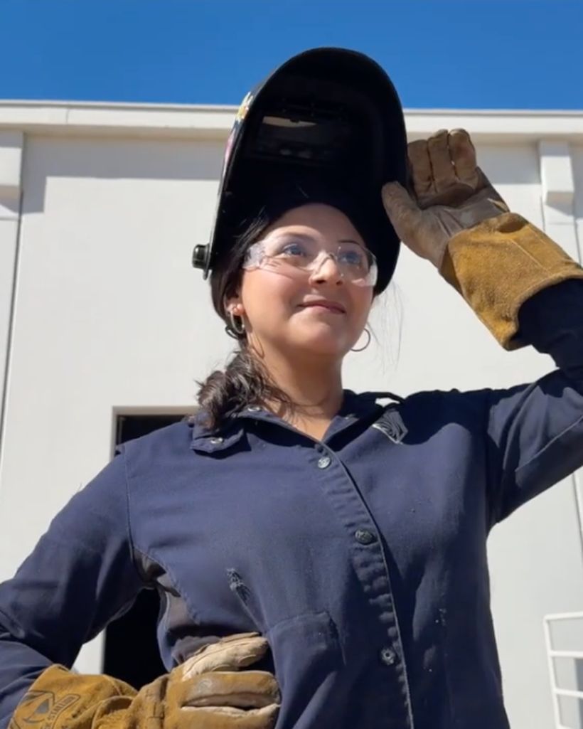 Wendy lifting her welding hood, in welding coveralls and gloves